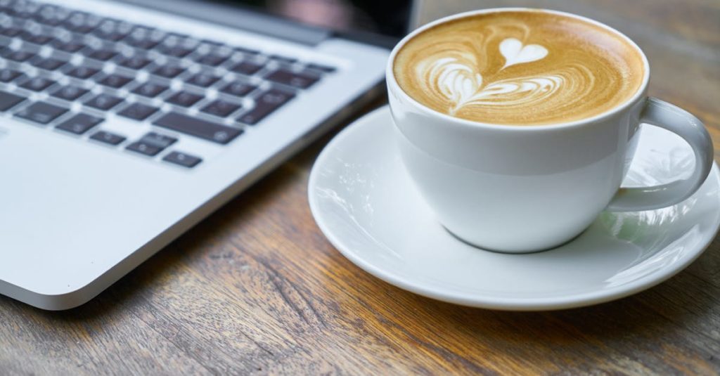 A cup of latte with intricate art beside a laptop on a wooden table, depicting a cozy work setting.
